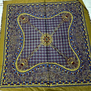 Puede incluir: Textil cuadrado con un patrón intrincado. El diseño central presenta una disposición geométrica de formas azules, burdeos y amarillas. El borde es de color verde oliva liso y los bordes tienen flecos. El textil es probablemente un artículo decorativo.
