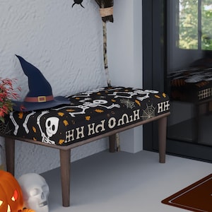 Puede incluir: Una entrada temática de Halloween con un banco, calabazas y decoraciones. El banco tiene un patrón de calaveras en blanco y negro. Hay un sombrero de bruja y una escoba. Murciélagos y arañas cuelgan de la pared. Una alfombra de bienvenida está en el suelo.