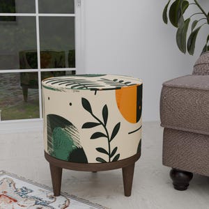 Może przedstawiać: Okrągły, kremowy puf z abstrakcyjnym wzorem kwiatowym w kolorze zielonym, pomarańczowym i czarnym. Puf ma cztery drewniane nogi.