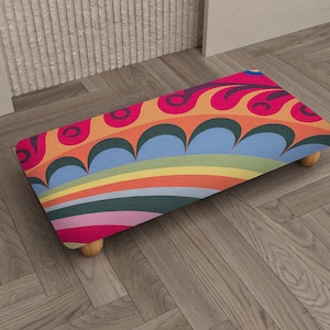 May include: A rectangular footstool with a vibrant, colorful design. The top features a pattern of swirling shapes in red, orange, and pink, with blue and green accents. It rests on four small, rounded wooden legs.