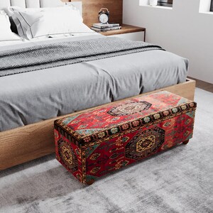 Op de afbeelding: Een rechthoekige opbergottoman met een rood patroon zit aan het voeteneinde van een bed. De ottoman heeft een gedetailleerd ontwerp in vintage stijl in rood, bruin en goud. Het bed heeft een licht houten frame en grijs beddengoed. De ottoman is ongeveer 100 cm lang.