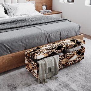 May include: A rectangular storage bench with a leopard print and gold chain design, open to reveal folded blankets and a gray scarf. The bench is positioned at the foot of a bed with a gray comforter.