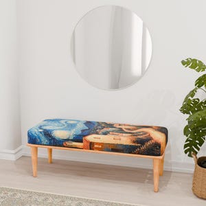 Puede incluir: Un banco de madera con un cojín estampado en azul y amarillo que representa una pintura de una noche estrellada. El banco tiene un diseño sencillo con cuatro patas y un asiento rectangular.