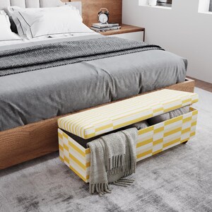 May include: A yellow and white striped storage bench with a hinged lid sits at the foot of a bed. The bench is open, revealing folded blankets and a gray scarf. The bed has gray bedding and white pillows.