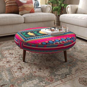 May include: A round upholstered ottoman with a vibrant, multi-colored geometric and floral pattern in shades of pink, blue, yellow, and black. The piece features short, tapered wooden legs and a flat top surface holding a book and glasses.