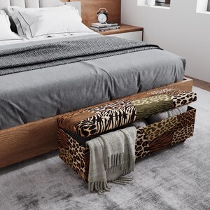 May include: A wooden storage bench with a patchwork animal print design sits at the foot of a bed. The bench is open, revealing folded blankets and a gray scarf. The bed has a gray comforter and white pillows.