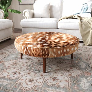 May include: A round ottoman featuring a brown and white faux fur upholstery with a spotted pattern. The piece is supported by four tapered wooden legs in a medium brown finish, providing a stable base for this low-profile furniture item.