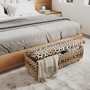 May include: A rectangular storage bench upholstered in a tan and black leopard print fabric. The bench features a hinged lid that is partially open, revealing a hollow interior. A light gray fringed throw blanket is draped over the front side.