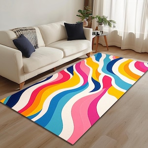 May include: A colourful abstract rug with wavy lines in shades of pink, blue, yellow, and orange on a white background. The rug is laid out on a wooden floor in front of a white sofa.