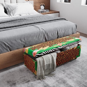 May include: A decorative storage bench with a vibrant leopard print design. The bench features a mix of patterns, including zebra stripes, green accents, and a leopard print. A gray blanket and pillows are visible inside the bench, and a gray scarf is draped over the edge.