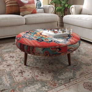 May include: A round ottoman featuring a vibrant, multi-colored pattern with prominent shades of red, teal, and orange. The piece is supported by four tapered wooden legs and holds a white book topped with black-rimmed eyeglasses and a glass.