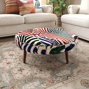 May include: A round ottoman featuring a vibrant, multi-coloured leaf pattern in shades of red, green, blue, and orange on a cream background. The piece is supported by four tapered wooden legs and has a piped edge along the top circular cushion.
