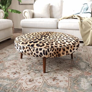 Puede incluir: Un taburete redondo con estampado de leopardo en tonos negro, marrón y crema. La pieza se apoya sobre cuatro patas de madera cónicas en un acabado marrón medio, con un detalle de ribete a lo largo del borde de la superficie circular.