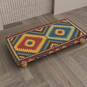 May include: A rectangular footstool with a colorful geometric pattern in red, yellow, blue, and green. The footstool is supported by four small, rounded wooden legs. The design features diamond and zigzag shapes, creating a vibrant, patterned surface.