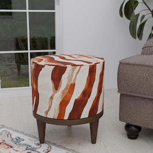 May include: A round, brown and white zebra-print ottoman with wooden legs. The ottoman is sitting on a white floor with a rug in the foreground.