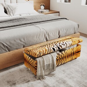 May include: A rectangular storage bench featuring a vibrant orange and black tiger stripe pattern. The lid is partially open, revealing an interior space. A light gray fringed throw blanket is draped over the front side of the upholstered bench.