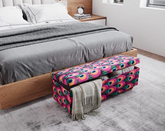 May include: A rectangular storage bench with a vibrant peacock feather pattern in pink, yellow, and teal. The bench is open, revealing folded blankets and a gray scarf. The bench is placed at the foot of a bed.