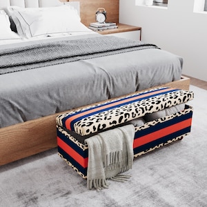 May include: A rectangular storage bench with a leopard print lid and navy blue and orange stripes. The bench is open, revealing folded blankets and a gray scarf. The bench is in front of a bed with gray bedding.