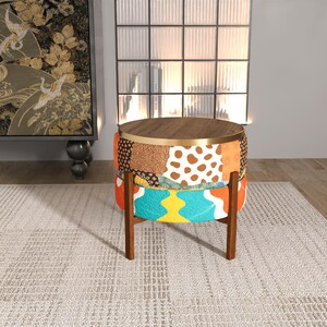 Puede incluir: Un otomán redondo y estampado con una tapa y patas de madera. La tela presenta un diseño de patchwork con varios patrones y colores, incluyendo marrón, naranja y turquesa. Las patas son de color marrón oscuro.