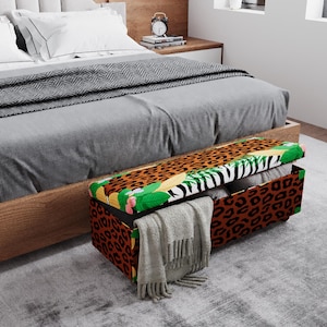 May include: A rectangular storage ottoman with a leopard print design on the sides and top. The top features a zebra stripe and tropical leaf pattern. A gray blanket is draped over the edge.