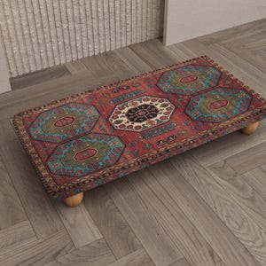 May include: A rectangular footstool with a red, teal, and beige patterned rug top. The rug design features geometric shapes and floral motifs. The footstool is supported by four small, round, wooden legs.