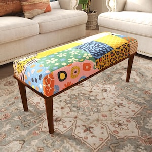 Peut inclure: Un banc ottoman rectangulaire doté de quatre pieds en bois brun foncé et d'un dessus rembourré recouvert d'un tissu patchwork vibrant. Le tissu présente une variété de motifs colorés, notamment des fleurs, des taches de léopard et des formes abstraites.
