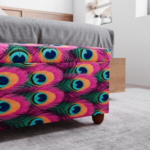a colorful, patterned ottoman or storage box with a vibrant design of peacock feathers in shades of pink, blue, green, and orange. The ottoman is placed on a gray carpeted floor, and there is a wooden cabinet visible in the background.