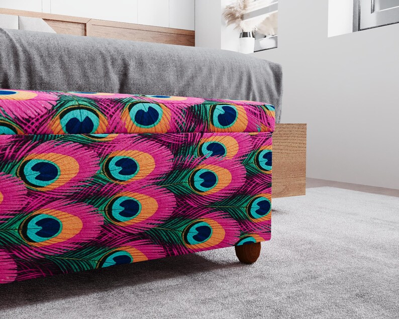 a colorful, patterned ottoman or storage box with a vibrant design of peacock feathers in shades of pink, blue, green, and orange. The ottoman is placed on a gray carpeted floor, and there is a wooden cabinet visible in the background.
