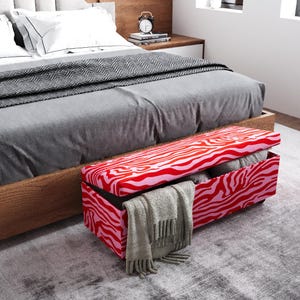 May include: A red and pink zebra-striped storage bench with the lid open, revealing folded blankets and pillows. A gray blanket with fringe hangs over the edge. The bench is positioned at the foot of a bed with gray bedding.