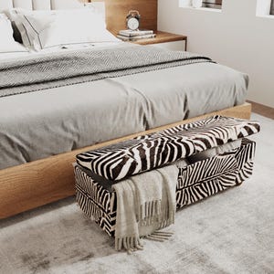 May include: A rectangular storage bench upholstered in a black and white zebra print fabric. The bench features a hinged lid that is partially open, revealing an interior space. A light beige fringed throw blanket is draped over the front side.