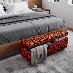 May include: A rectangular storage bench with a leopard print design in red and black sits at the foot of a bed. The bench is open, revealing pillows and a gray blanket. The bed has a gray comforter and a wooden frame.