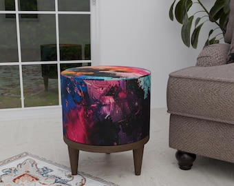 Puede incluir: Un otomano redondo de colores con un diseño abstracto negro, rosa, azul y morado. El otomano tiene cuatro patas de madera.