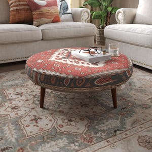 May include: A round ottoman featuring a red, white, and blue patterned fabric with a geometric design. The piece is supported by four dark brown wooden legs and holds a white book topped with black-rimmed eyeglasses and a clear glass.