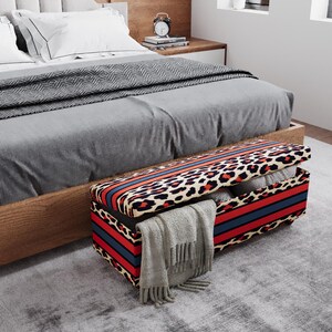 May include: A rectangular storage bench with a leopard print design, accented with red and blue stripes. The bench is open, revealing folded blankets and a gray scarf. The bench is placed at the foot of a bed with gray bedding.