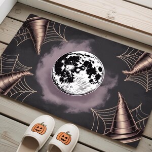 May include: A rectangular Halloween doormat with a dark background, featuring a full moon, witch hats, and spiderwebs. Two pairs of white slippers with jack-o'-lantern faces are at the bottom. The design is in shades of black, purple, and brown.