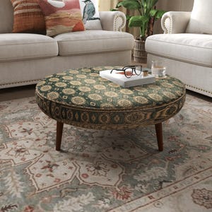 Puede incluir: Una mesa de centro tipo otomana redonda con una superficie de tela verde oscuro y un patrón geométrico beige repetitivo. Los laterales están tapizados con tela de estampado de veta de madera oscura. Se apoya sobre cuatro patas cónicas de madera marrón.