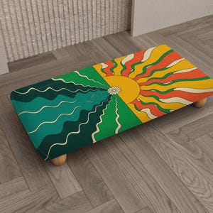 Puede incluir: Un reposapiés rectangular con un diseño retro que presenta un motivo de sol y océano. La parte superior está dividida en cuadrantes verdes, turquesa, amarillos y naranjas. El taburete está sostenido por cuatro patas de madera pequeñas y redondeadas.