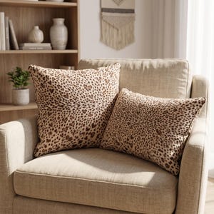 May include: Two square throw pillows featuring a brown and black leopard print pattern. The pillows are made of fabric and have a soft, plush appearance. The leopard spots are arranged in a dense, repeating design across the entire surface.