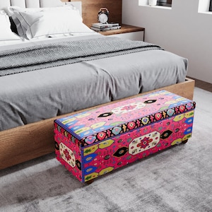 Könnte beinhalten: Eine rosa Truhe mit farbenfrohem Blumenmuster steht am Fuß eines Bettes. Die Truhe hat eine rechteckige Form mit einem Klappdeckel und Holzbeinen. Das Bett hat einen Holzrahmen, graue Bettwäsche und weiße Kissen.