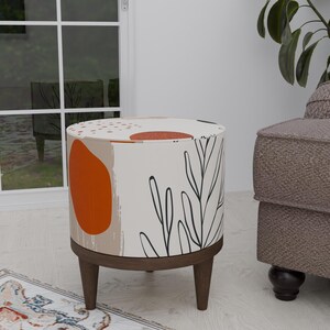 May include: A round, white ottoman with a brown wooden base and legs. The ottoman is covered in a fabric with an abstract pattern of orange, brown, and black. The pattern features leaves and circles.