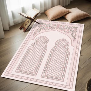 Puede incluir: Una alfombra de oración rosa claro con un diseño floral y dos patrones en forma de arco. La alfombra tiene un borde con pequeños motivos florales repetidos. Un atril de madera con un libro abierto es visible en el fondo.