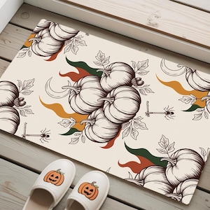 Könnte beinhalten: Eine rechteckige Fußmatte mit cremefarbenem Hintergrund zeigt ein sich wiederholendes Muster aus Kürbissen, Blättern und Spinnen im Vintage-Stil. Zwei weiße Hausschuhe mit Kürbisgesichtern sind im Vordergrund.