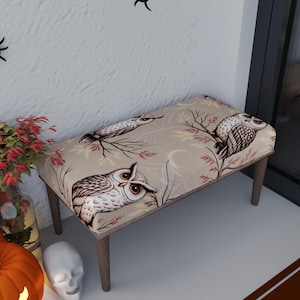 May include: A small bench with a beige owl print. The bench has four wooden legs and a rectangular seat. The owl print features brown and white owls, branches, and spiderwebs. The bench is a decorative piece.