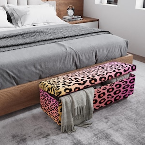 May include: A rectangular storage bench with a leopard print design in shades of pink, yellow, and black. The bench is open, revealing a gray blanket inside. The bench is at the foot of a bed with gray bedding.