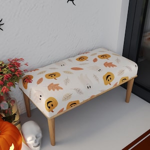 May include: A small wooden bench with a Halloween-themed design. The bench features a white fabric top adorned with orange jack-o'-lanterns, autumn leaves, and ghost illustrations. The bench has four wooden legs and is suitable for indoor use.