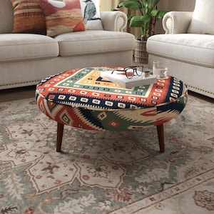 May include: A round ottoman featuring a vibrant, multi-colored geometric pattern in shades of red, blue, green, and cream. The piece is supported by four short, dark brown wooden legs and is topped with a white book and black-rimmed glasses.