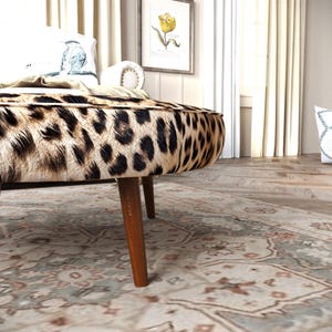 a leopard print ottoman with a wooden base, placed on a patterned rug in a room with a window and curtains.