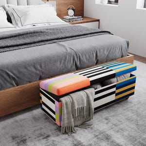 May include: A colorful storage bench with a striped and geometric design, open to reveal folded blankets and pillows. The bench has a rainbow-colored top and is placed at the foot of a bed with gray bedding.