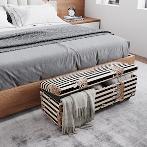 May include: A storage ottoman with a black and white striped pattern and leopard print accents. The ottoman is open, revealing pillows and a folded blanket. The ottoman is placed at the foot of a bed with gray bedding.