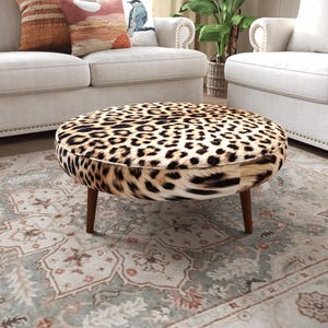 a living room with a leopard print ottoman placed on a patterned rug. The room is furnished with two white couches and a potted plant, creating a cozy and stylish atmosphere.
